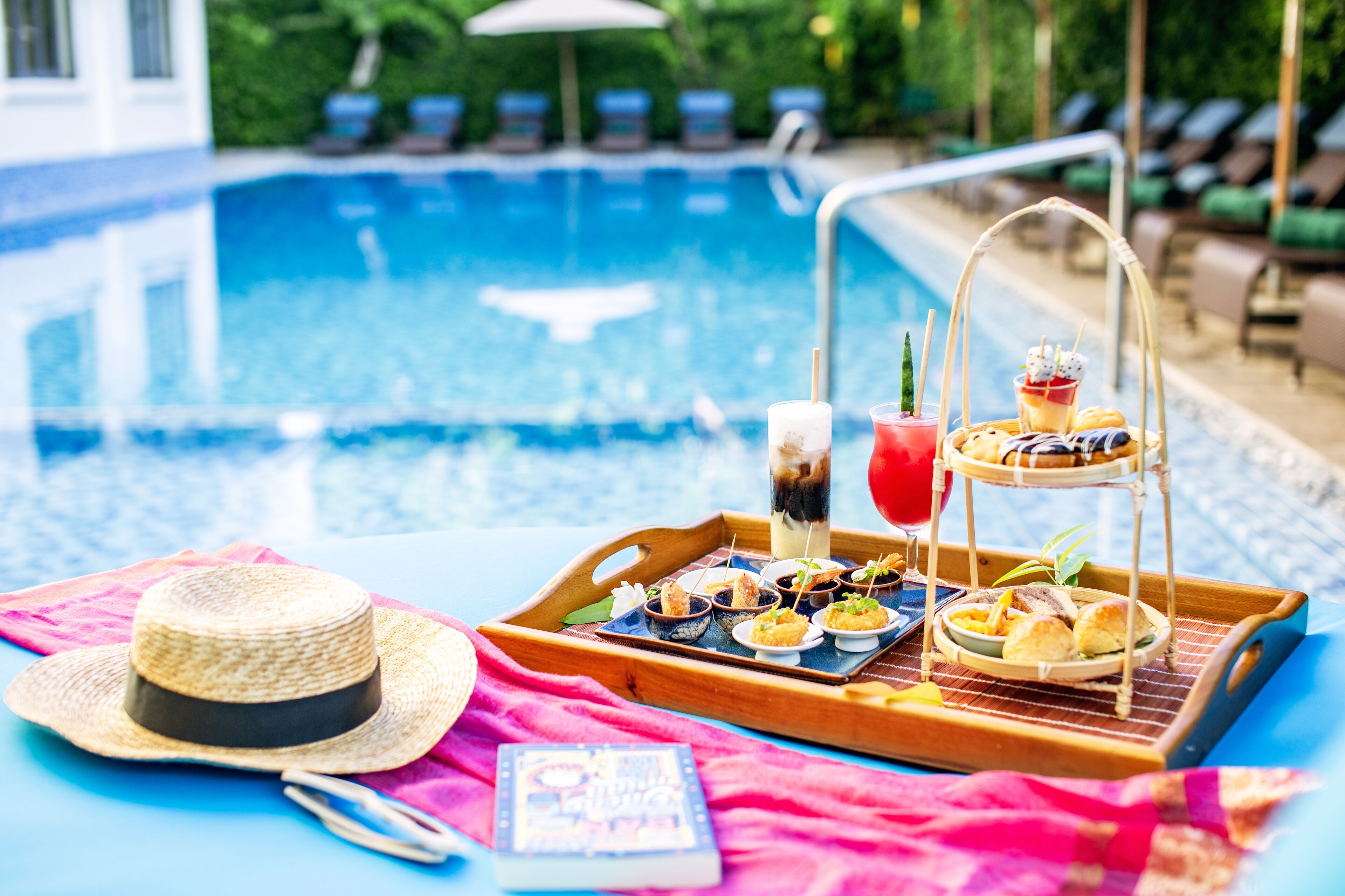 A poolside setting with a tray of food and drinks, a straw hat, and a book on a pink towel.