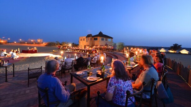 A dining area with tables and chairs, a building in the background, and a body of water. - Raisar Camp (Bikaner)