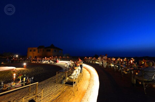 An outdoor dining area with tables and chairs, a building in the background, and a clear sky.