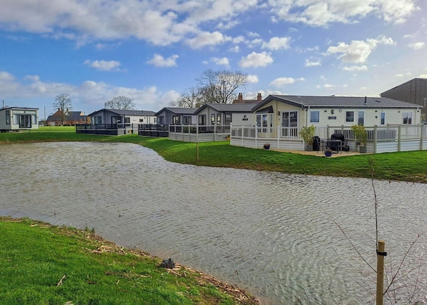 A residential area with houses along a water body, featuring a grassy lawn and a paved walkway. - Platinum 4 berth Lodge (Helsey)