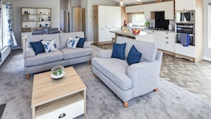 Un soggiorno moderno con un divano grigio, un tavolino in legno e una zona cucina con mobili e elettrodomestici bianchi.