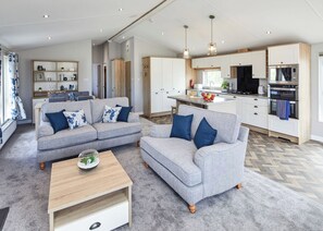 A modern living room with a grey sofa set, wooden coffee table, and a kitchen area with white cabinets and appliances.