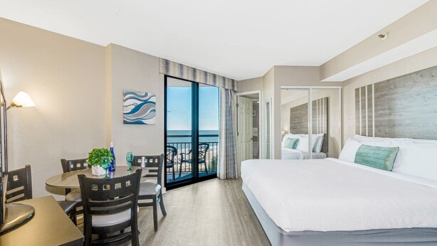 Une chambre d’hôtel avec un grand lit, un coin repas avec des chaises et une table, une vue sur la mer par une porte vitrée et un tableau au mur.