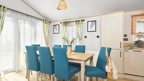 A dining area with a wooden table and blue chairs, a hanging light fixture, and a kitchen area with cabinets and a window.