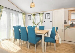 Ein Essbereich mit Holztisch und blauen Stühlen, einer hängenden Leuchte sowie eine Küchenzeile mit Schränken und einem Fenster.