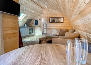 A cozy cabin interior with a wooden ceiling, a sofa, a table with glasses, and a kitchen area.