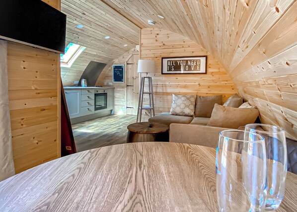 A cozy cabin interior with a wooden ceiling, a sofa, a table with glasses, and a kitchen area.