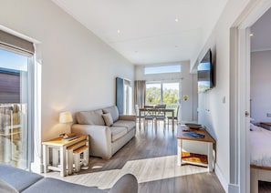 A modern living room with a sofa, dining table, and chairs, a TV mounted on the wall, and a large sliding glass door leading to a balcony.