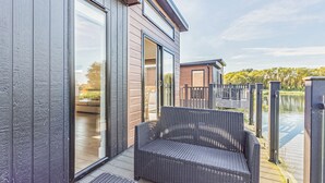 A modern building with large sliding glass doors, a wicker chair, and a wooden deck overlooking a body of water. - 3 bedroom accommodation in Poulton-Le-Fylde (Poulton-Le-Fylde)