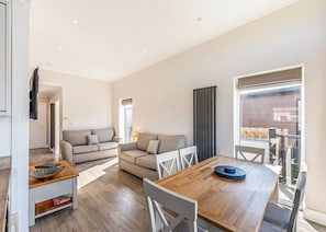 A modern living room with a sofa, dining table, and chairs. There is a window with curtains, a door, and a view of the outside.