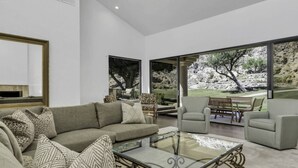 A modern living room with a large glass coffee table, a grey sofa, and two armchairs. There is a large window providing a view of a landscaped garden. - Premium PGA West Location Large Saltwater Pool/Spa Fire Pit Spectacular Mtn Views: #067312 (La Quinta)