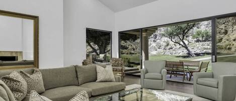 A modern living room with a large glass coffee table, a grey sofa, and two armchairs. There is a large window providing a view of a landscaped garden.