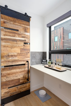 A modern kitchen with a wooden wall panel, a window with a view of a brick building, and a countertop with a wine sign and glassware.