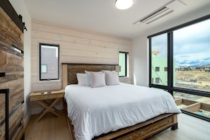 A bedroom with a large bed, wooden headboard, a desk, and a view of a construction site through large windows.