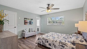 A bedroom with a bed, a gray sofa, a ceiling fan, a window with blinds, and a painting on the wall.
