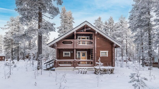 Kaksikerroksinen hirsihökkelö, jossa on parveke, ja sen ympärillä on lumipeitteisiä puita.