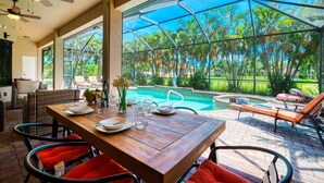 A dining area with a wooden table, red cushioned chairs, and a view of a swimming pool and tropical garden.