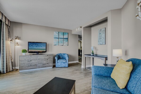 A modern living room with a flat-screen TV, a blue armchair, a sofa with a patterned pillow, a wooden coffee table, and a dining area with a table and chairs.