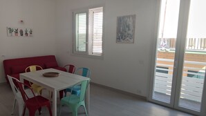A dining area with a white table and colorful chairs, a red sofa, and a window with blinds. - Donna Beatrice AmMare beach house (Donnalucata)