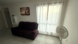 Ein Wohnzimmer mit einer Couch, einem Standventilator, einem Fenster mit Vorhängen und einem an der Wand hängenden Bild.