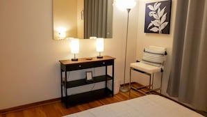 Ein Hotelzimmer mit einem Holztischchen, einem Spiegel, einer Stehlampe, einem Sessel und einem gerahmten Bild an der Wand.