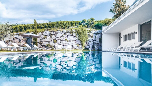 A swimming pool with lounge chairs, a stone wall, and a building with large windows. - Belvilla by OYO Palm Loggia (Meran)