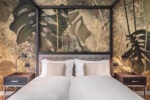 Uma cama de casal com cabeceira estofada, duas mesas de cabeceira com abajures, e uma parede com padrão de folhas tropicais.
