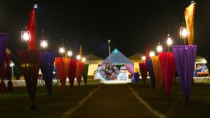 Un espace aménagé avec des tentes, des rideaux et des guirlandes lumineuses de couleurs vives, une scène où se produisent des artistes, et une foule de spectateurs.