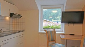 Una piccola cucina con mobili bianchi, una finestra con vista su una casa e alberi, una TV a parete e due sedie a righe a un tavolo di legno.