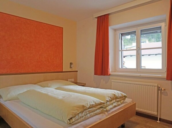 A single bed with a wooden headboard, a window with orange curtains, and a radiator below.