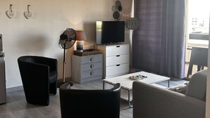 A living room with a TV, a fan, a lamp, and a sofa. - 3-room apartment with balcony and parking, pool access, sports areas (Le Barcarès)