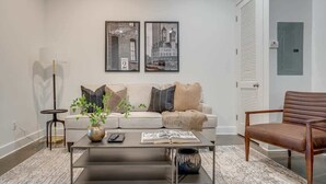 A modern living room with a sofa, coffee table, and a chair. There are two framed pictures on the wall, a plant on the coffee table, and a door with a sign. - Lofts at 30th-Urban Nest-4 Mins to Broadway! (Nashville)