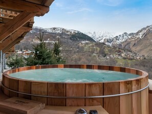 Ein hölzernes Whirlpool mit Metallgeländer, mit Blick auf ein dorfähnliches Bergdorf mit schneebedeckten Gipfeln.