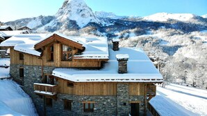 Een chalet van hout met meerdere verdiepingen, stenen muren, sneeuwbedekt dak en balkons, gelegen tegen een bergachtige achtergrond.