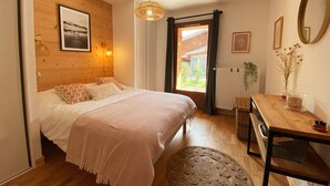 A bedroom with a bed, a wooden headboard, a hanging light fixture, a window with curtains, a radiator, a small table with a vase, and a mirror on the wall. - Luxurious Mountain Apartment, 2 Beds, Terrace + High-End Amenities, Close to Village & Ski Bus (Samoëns)