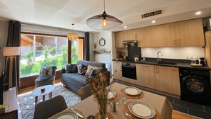 A modern kitchen with wooden cabinets, a dining area with a table set for two, and a living room with a sofa and a view of the outdoors. - Luxurious Mountain Apartment, 2 Beds, Terrace + High-End Amenities, Close to Village & Ski Bus (Samoëns)
