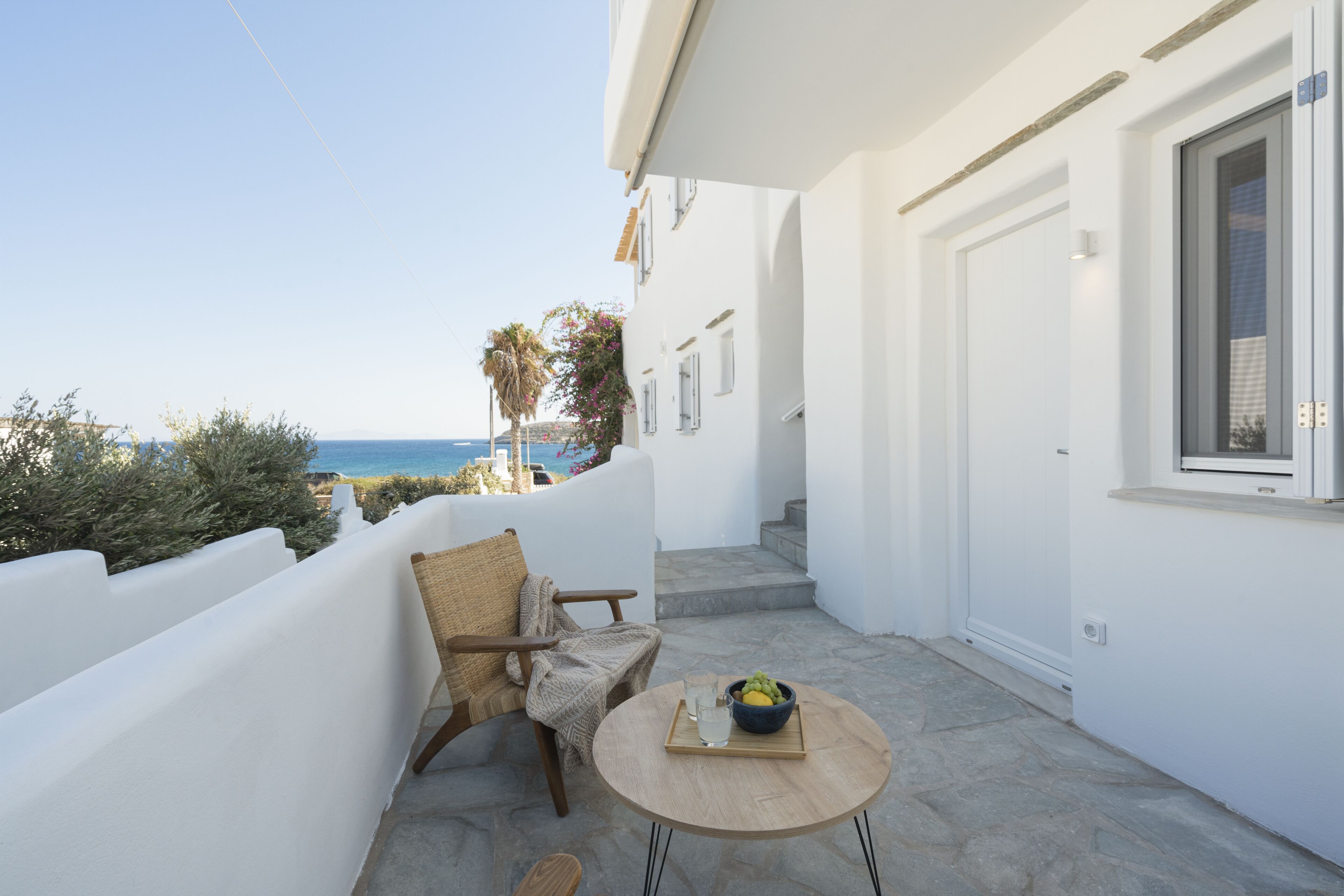 A balcony with a view of the sea, a wicker chair, a small table with a bowl of fruit, and a white building with a door and window.