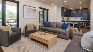 A modern living room with a sofa, armchairs, and a coffee table. A kitchen is visible in the background with dark cabinets and a dining area. - Luxury Condo 5 Blocks from Downtown (Ketchum)