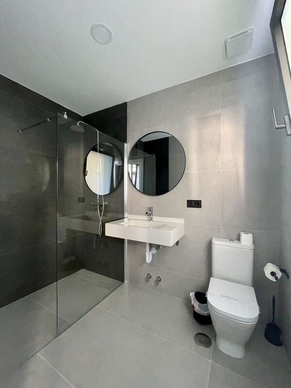 Un baño moderno con cabina de ducha de vidrio, un lavabo blanco, un inodoro y un espejo redondo.
