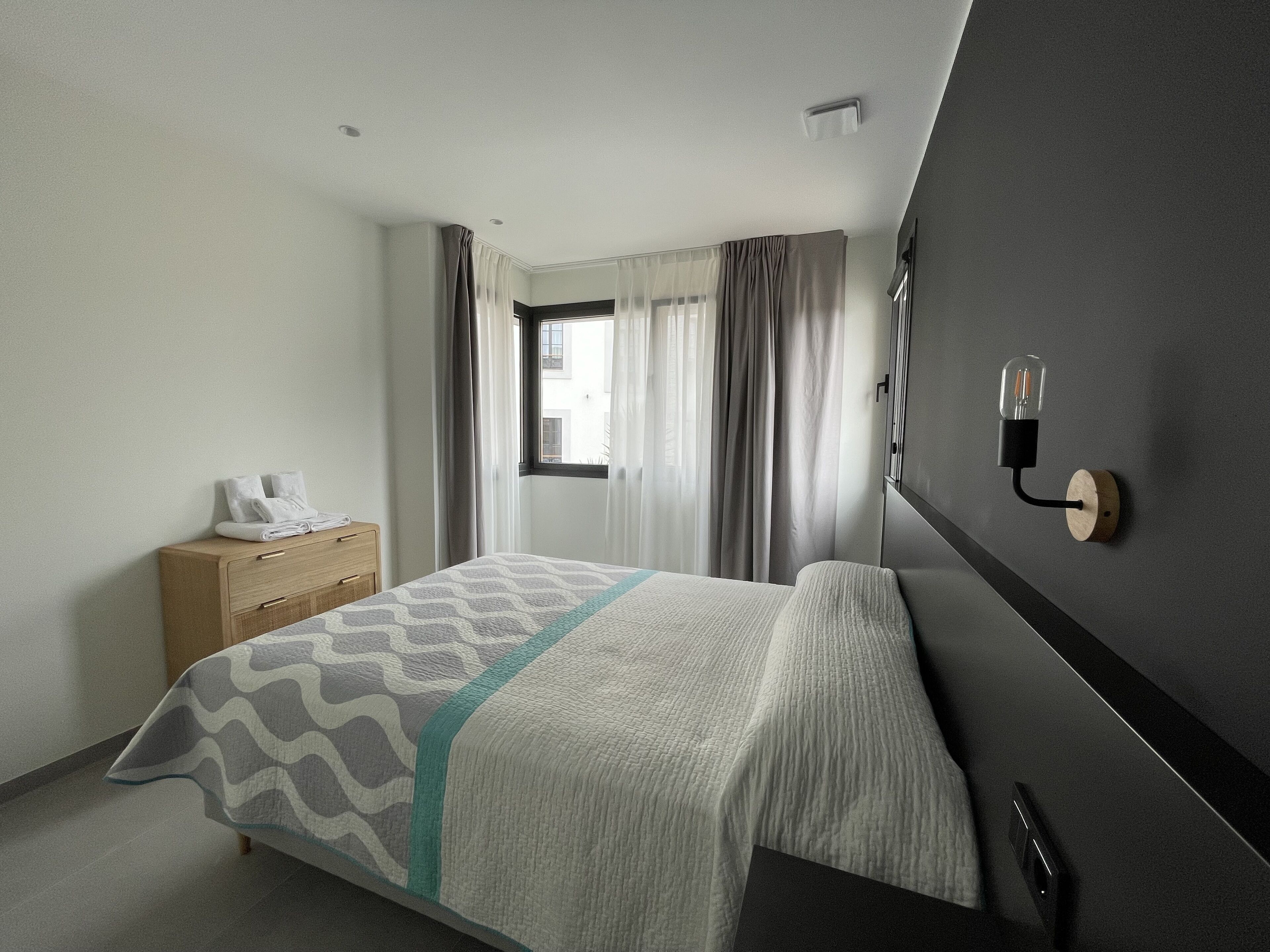 A modern hotel room with a neatly made bed, a wooden dresser, and a wall-mounted light fixture.