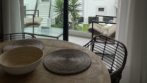 A dining area with a glass table, woven placemats, and wicker chairs. A view of a building with balconies and a palm tree is visible through the window. - Apartamentos Las Indianas (Llanes)
