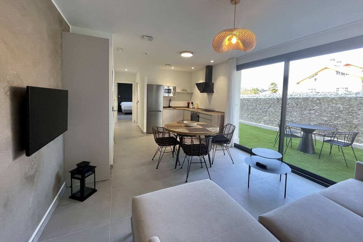A modern living room with a flat-screen TV mounted on the wall, a dining area with a table and chairs, and a kitchen with a microwave and refrigerator.
