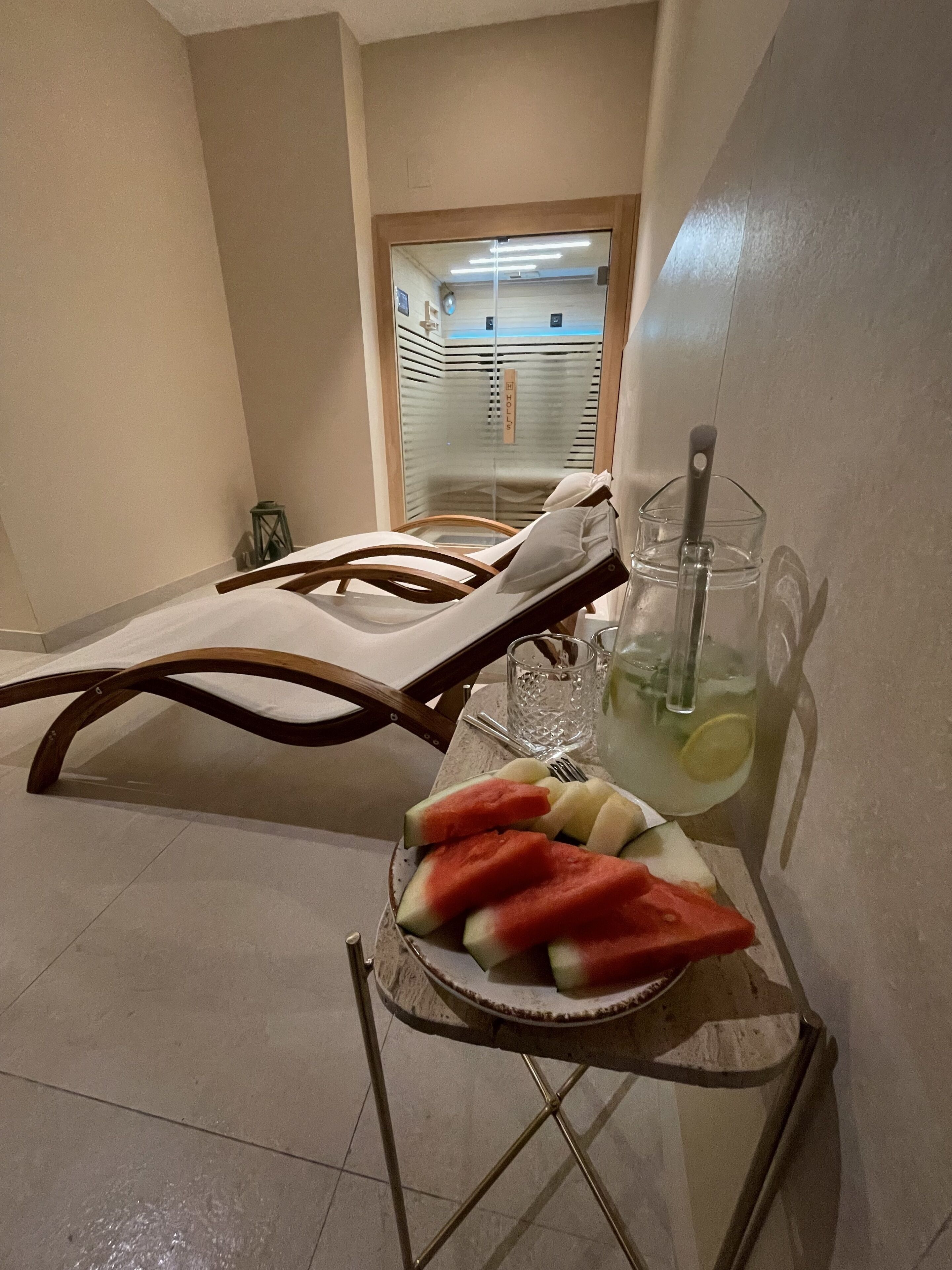 Una habitación con una silla de descanso, una mesa con un vaso de agua y un frutero, y vista a un pasillo con una puerta.