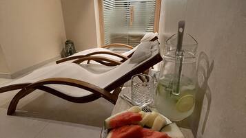 Una habitación con una silla de descanso, una mesa con un vaso de agua y un frutero, y vista a un pasillo con una puerta.