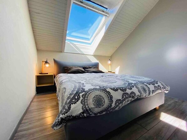Une chambre avec un puits de lumière, un lit avec une literie à motifs, deux lampes de chevet et un plancher en bois.
