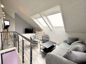 Un salon de style industriel, avec un plafond haut, des fenêtres de toit, un escalier, un téléviseur à écran plat, un canapé et une table basse.