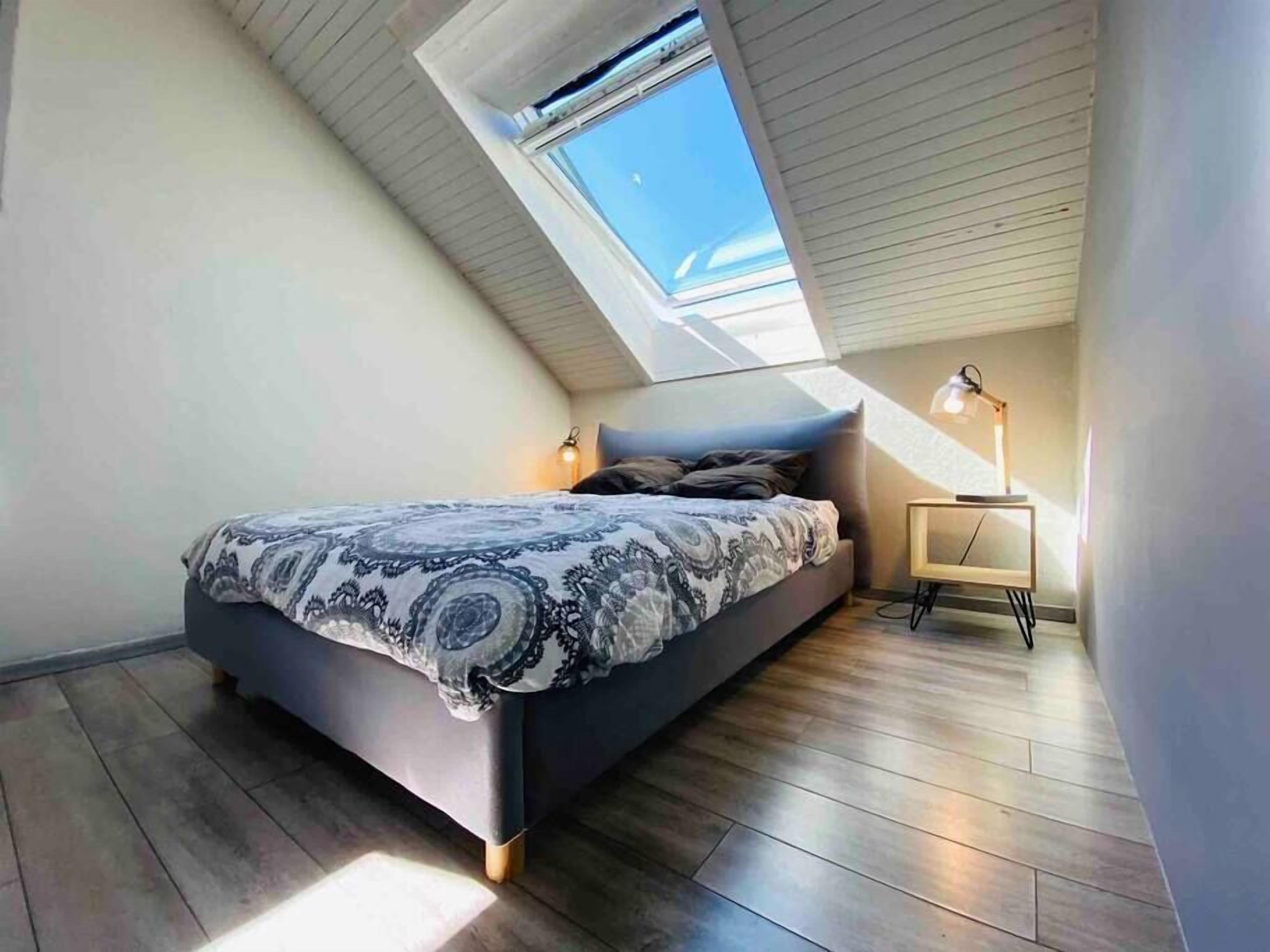 A bedroom with a sloped ceiling, a bed with patterned bedding, a bedside table with a lamp, and a view of the sky through a skylight.