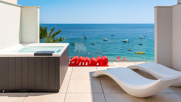 A balcony with a hot tub, lounge chairs, and a view of the sea with boats.
