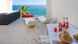 A terrace with a view of the sea, white lounge chairs, a table with drinks and snacks, and a bottle of liquor.