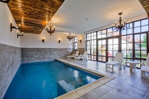 Une piscine intérieure dotée d’un plafond en bois, d’un lustre et offrant une vue sur l’extérieur grâce à de grandes fenêtres.